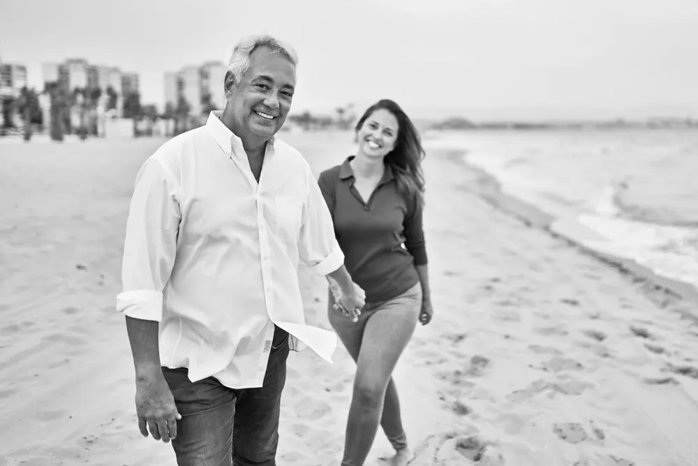 A gentelman in the foreground smiling and walking on the beach while a woman in the background smiles and walks behind him